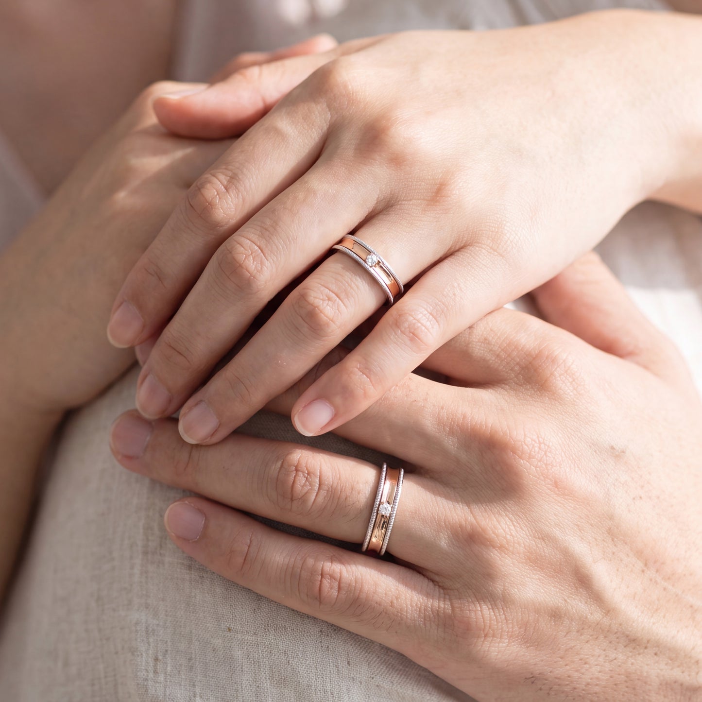 Two hands resting on a light fabric surface, each wearing a slim rose gold band ring with a small clear stone.