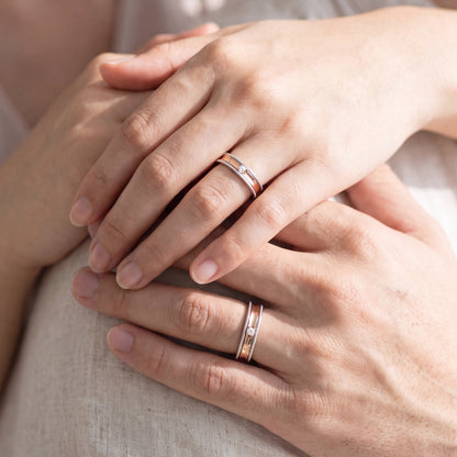 Two hands resting on a light fabric surface, each wearing a slim rose gold band ring with a small clear stone.